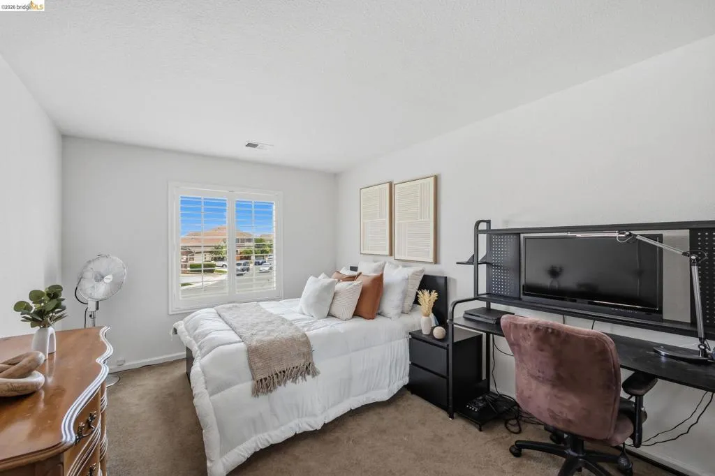 Carpeted bedroom featuring a white framed window with plantation shutters, light-toned walls, and a black desk with integrated shelving