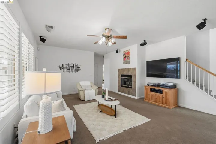 Living area featuring a fireplace with a stone-tile surround, white plantation shutters, a ceiling fan, carpet flooring, and a white-painted staircase with wood handrails