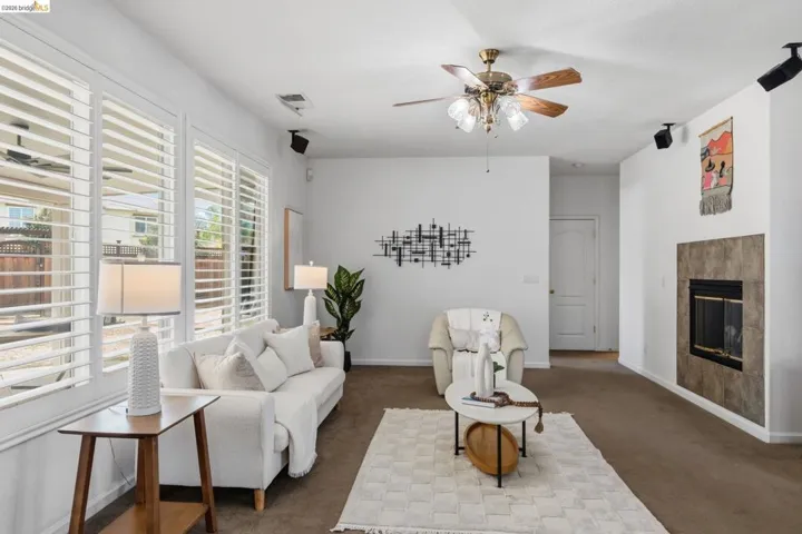 Spacious living area featuring extensive window panels with white louvered shutters, a ceiling fan with integrated lighting, and a fireplace with a tiled surround