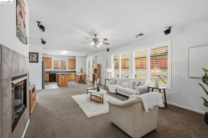 Spacious open-concept living area featuring a tile-surround fireplace, multiple windows with plantation shutters, ceiling fan, and carpeted flooring