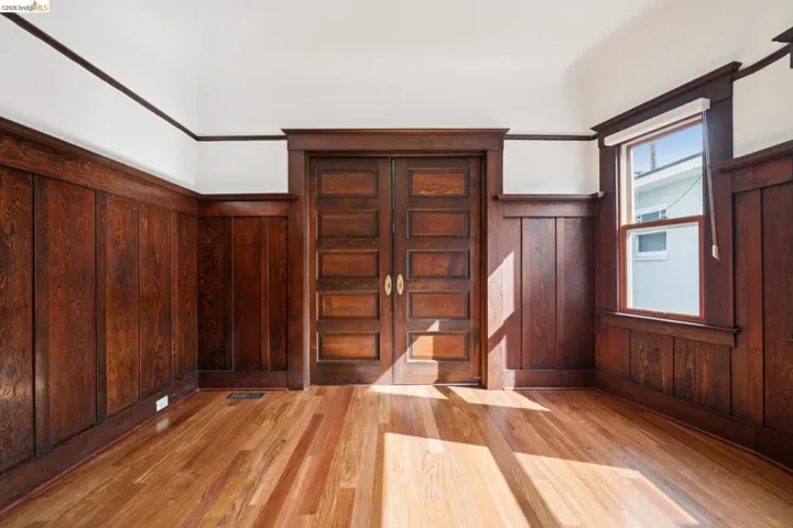 Spacious room featuring wood-finish flooring, wood paneling, wood trim, wood-paneled pocket doors with brass hardware, and a single window with a roller shade