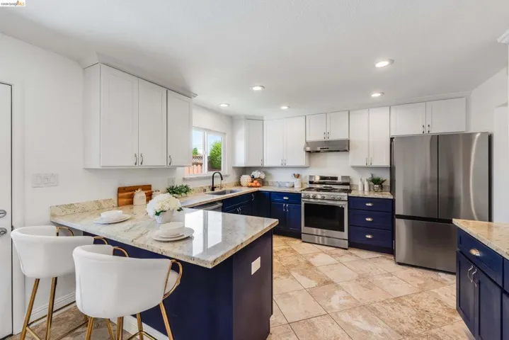 Kitchen featuring two-tone cabinetry with white upper and blue lower cabinets, stainless steel appliances, light-toned stone countertops, a breakfast bar, and recessed lighting