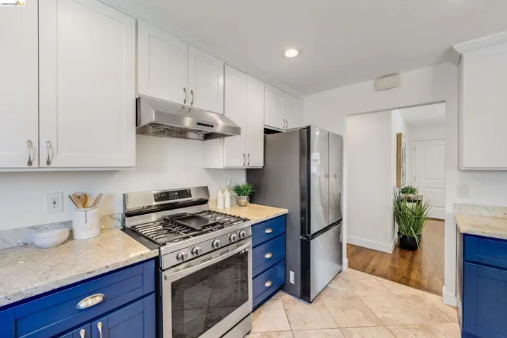 Kitchen featuring white upper cabinetry and blue lower cabinetry with silver hardware, light-colored stone countertops, and stainless steel appliances including a gas range and refrigerator