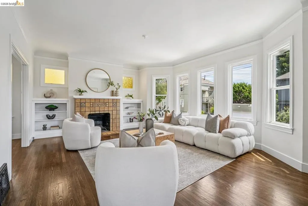 Living room featuring a tiled fireplace with mantel, built-in shelving, hardwood flooring, crown molding, and bay windows