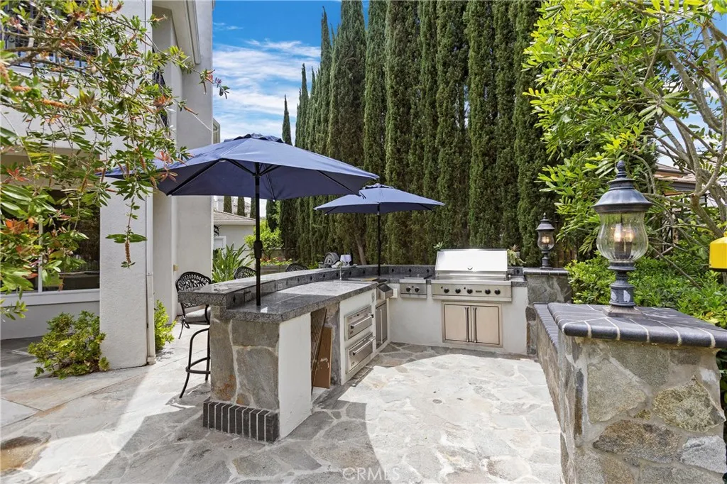 Discover this inviting outdoor kitchen space withViking BBQ, Cooktop, Warming Drawers, outdoor lighting and shady umbrellas, perfect for entertaining, surrounded by lush greenery and ample seating.