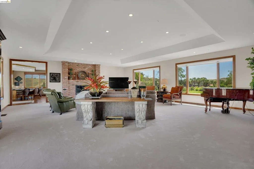 Spacious living area featuring expansive windows, a brick-faced fireplace, recessed lighting, a tray ceiling, and light-toned carpeting