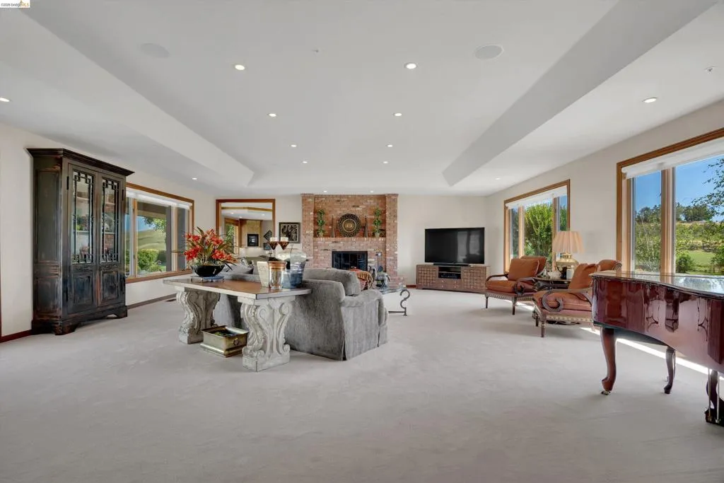Spacious living area featuring a prominent brick fireplace, light-toned carpeting, ample recessed lighting, and large windows with wooden frames offering exterior views