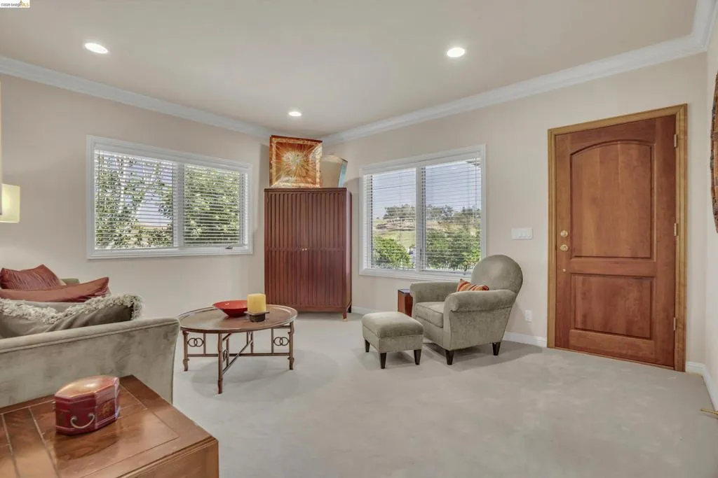 Carpeted living area featuring two large windows with blinds, crown molding, recessed lighting, and a solid wood door with an arched panel