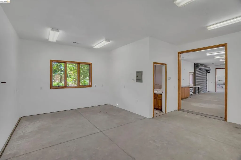 Spacious concrete-floored room featuring multiple fluorescent ceiling lights, a large wood-framed window, and a separate room with a vanity