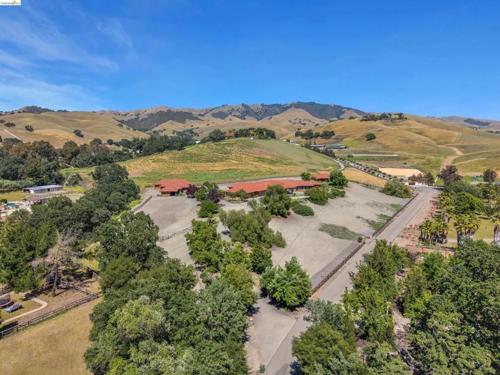 Expansive property featuring a Mediterranean-style residence with a terracotta tile roof, surrounded by mature trees, fenced paddocks, and distant rolling hills