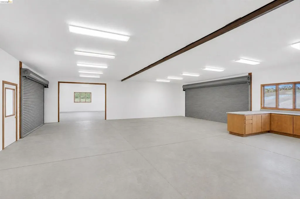Spacious multi-purpose room featuring concrete flooring, multiple roll-up doors, extensive overhead lighting, and built-in cabinetry with countertops