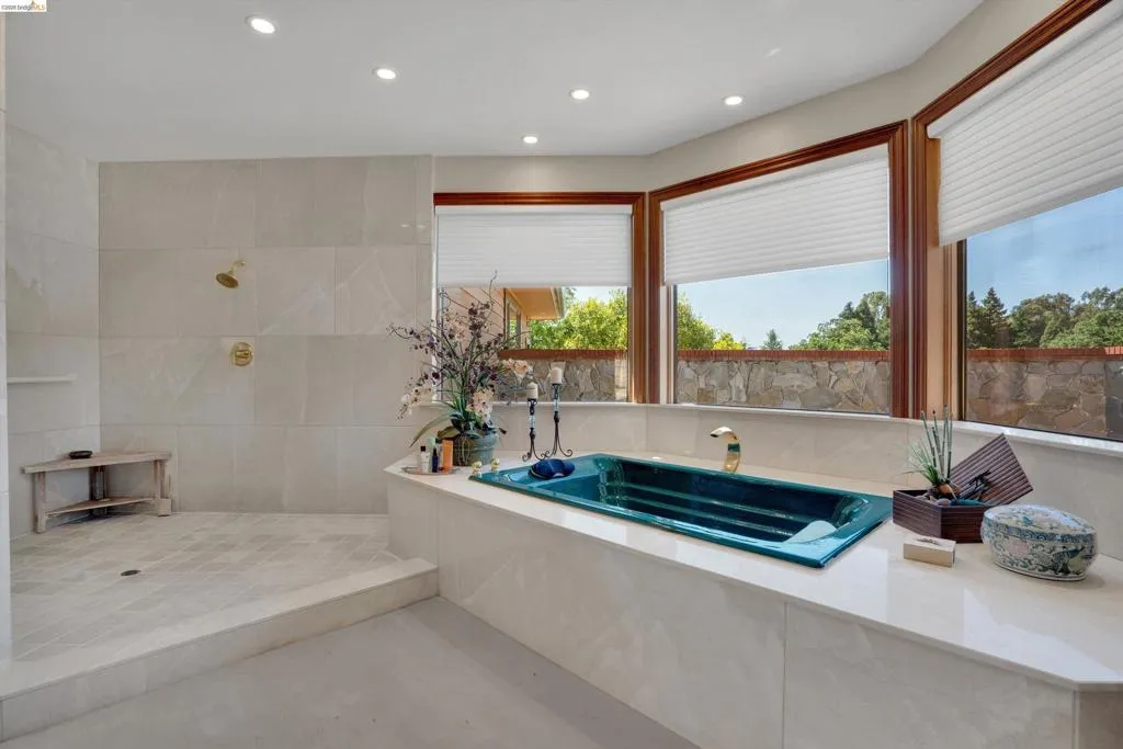 Bathroom featuring a teal soaking tub with brass fixtures, a walk-in shower with gold-tone hardware and built-in shelving, and large windows with wood-frame trim