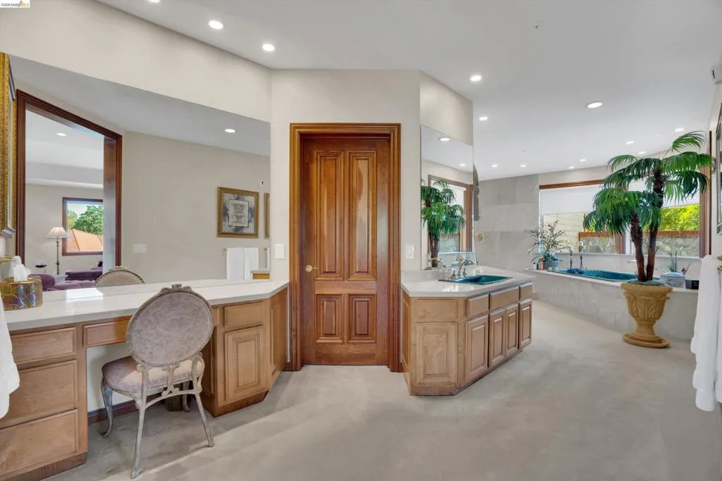 Spacious bathroom featuring a built-in vanity, light wood cabinetry, white countertops, dual sinks with chrome fixtures, and a large soaking tub