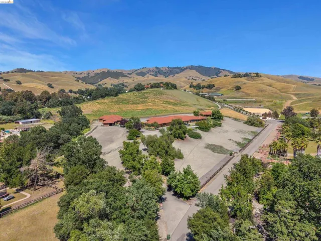 Expansive property featuring a Mediterranean-style residence with a terracotta tile roof, surrounded by mature trees, fenced paddocks, and distant rolling hills