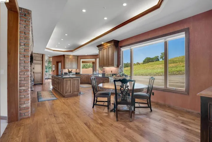 Spacious open-concept kitchen and dining area featuring wood-finish flooring, recessed lighting, a brick accent wall, and a large multi-pane window with a landscape backdrop