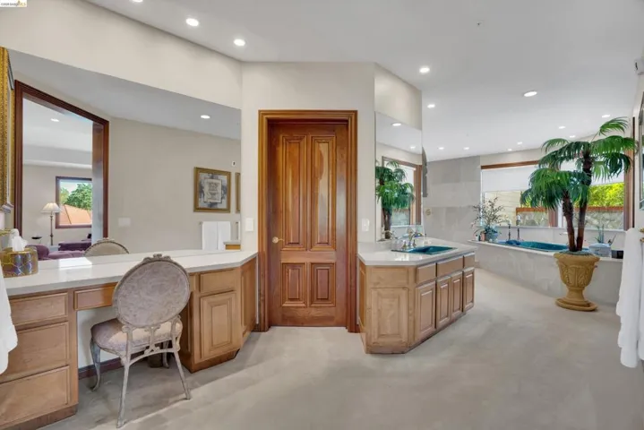 Spacious bathroom featuring a built-in vanity, light wood cabinetry, white countertops, dual sinks with chrome fixtures, and a large soaking tub