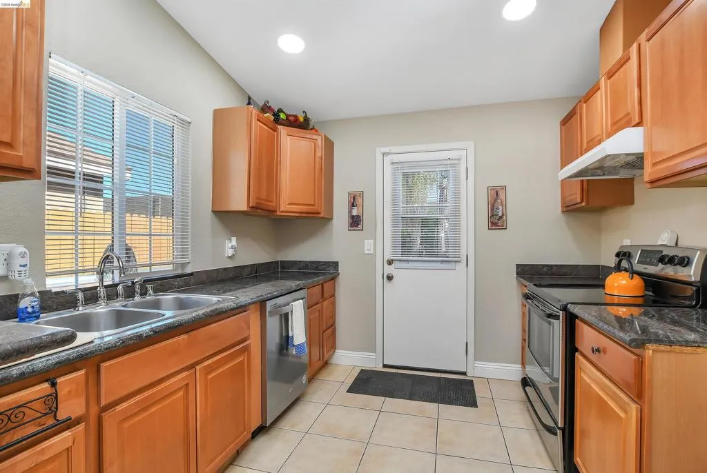 Kitchen featuring warm wood cabinetry, dark countertops, a double basin stainless steel sink, tile flooring, and a stainless steel appliance suite