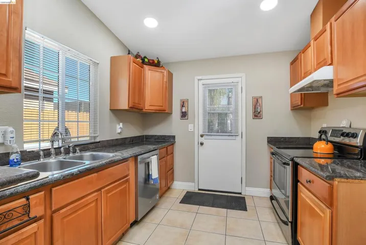 Kitchen featuring warm wood cabinetry, dark countertops, a double basin stainless steel sink, tile flooring, and a stainless steel appliance suite
