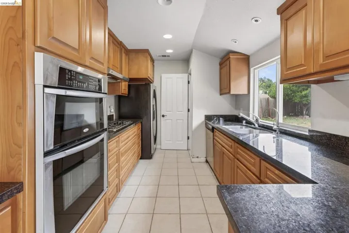 Galley kitchen featuring wood cabinetry, dark stone countertops, stainless steel double ovens, a gas cooktop, and a tile floor