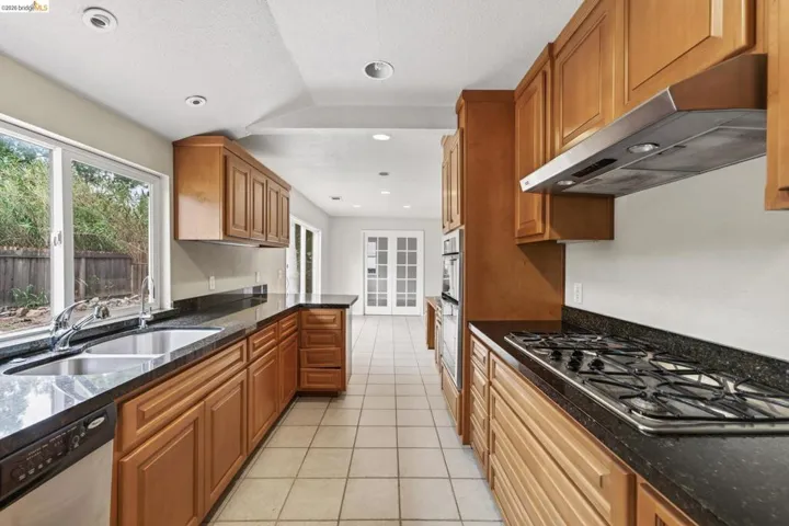 Kitchen featuring ample cabinetry, dark countertops, a gas range with stainless steel hood, built-in double ovens, and a dual basin sink with a window overlooking the property