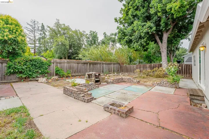 Expansive backyard featuring a combination of concrete and paved patio areas, mature trees, wood fencing, and brick-edged garden beds