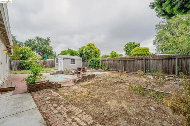 Expansive backyard featuring a gray storage shed, a wood fence, concrete patio areas, and brick hardscaping