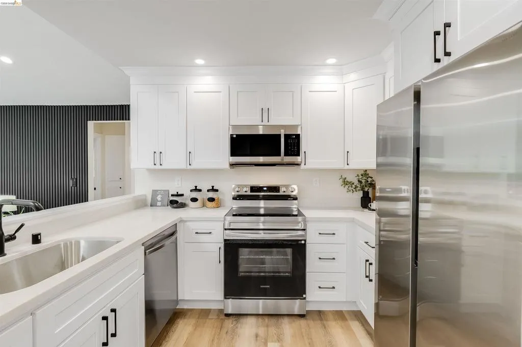 Bright kitchen featuring white shaker cabinetry, stainless steel appliances, light-toned countertops, a dual basin sink with a matte black faucet, and wood-finish flooring