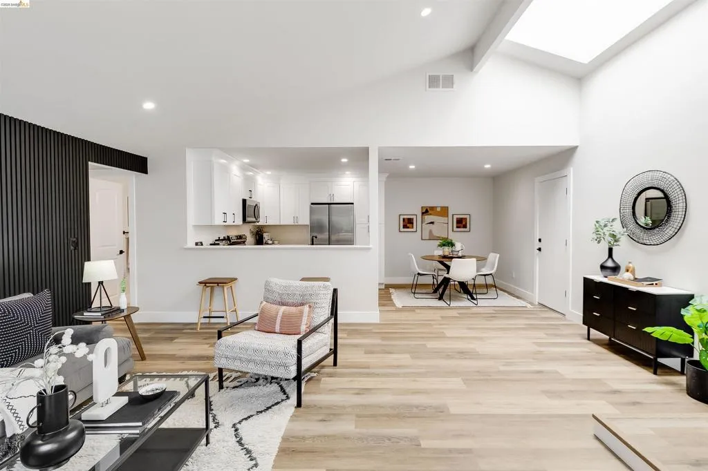 Open-concept living space featuring vaulted ceilings with skylights, light wood-finish flooring, a modern kitchen with white cabinetry and stainless steel appliances, and a dedicated dining area