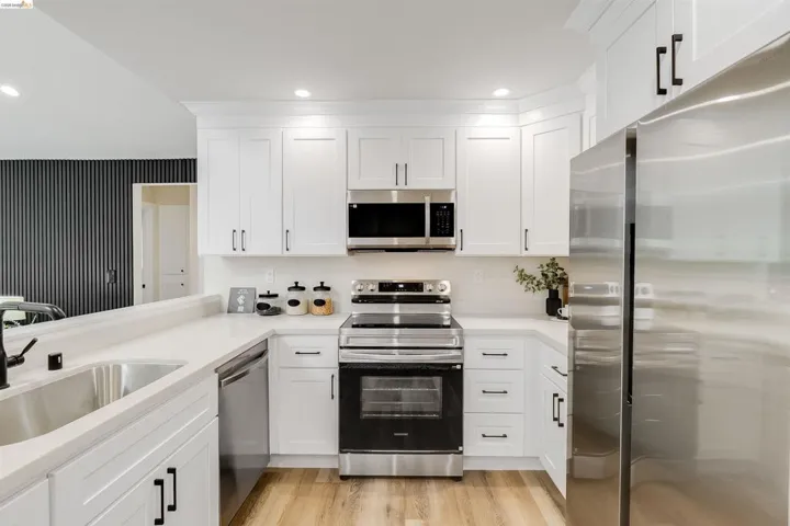 Bright kitchen featuring white shaker cabinetry, stainless steel appliances, light-toned countertops, a dual basin sink with a matte black faucet, and wood-finish flooring