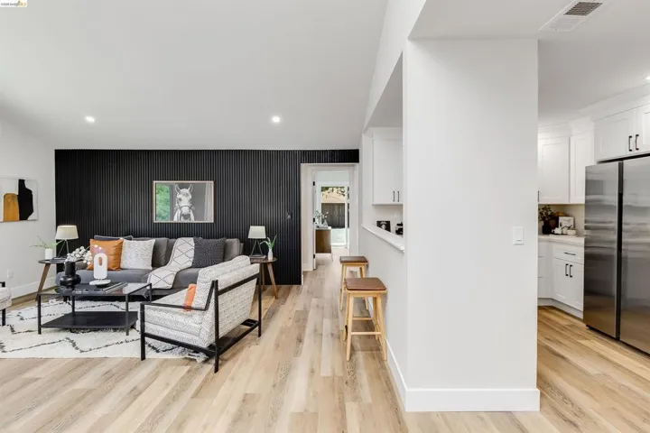 Bright interior featuring a striking black accent wall with vertical paneling, light wood-finish flooring, white kitchen cabinetry with dark hardware, recessed lighting, and a stainless steel refrigerator