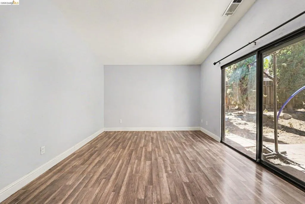 Spacious room featuring wood-finish flooring, light gray walls, white baseboards, and a large sliding glass door opening to an outdoor area with natural landscaping
