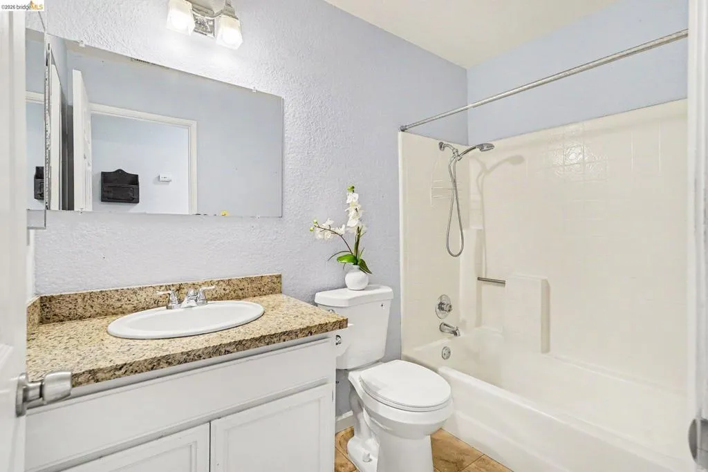 Bathroom featuring a white vanity with a speckled countertop, an oval undermount sink, a white toilet, and a combination shower-tub unit with a white tiled surround