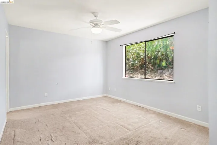 Carpeted room featuring light blue walls, a ceiling fan with integrated lighting, a large window with exterior foliage, and white baseboards