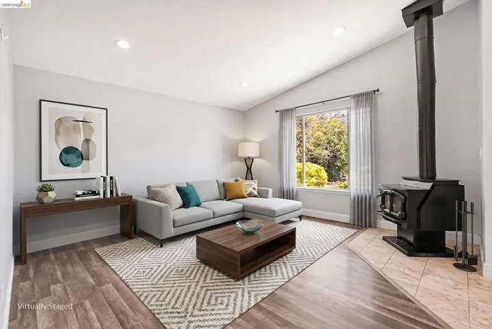 Living area featuring vaulted ceilings, recessed lighting, wood-finish flooring, a large window, and a built-in wood-burning stove with a tiled hearth
