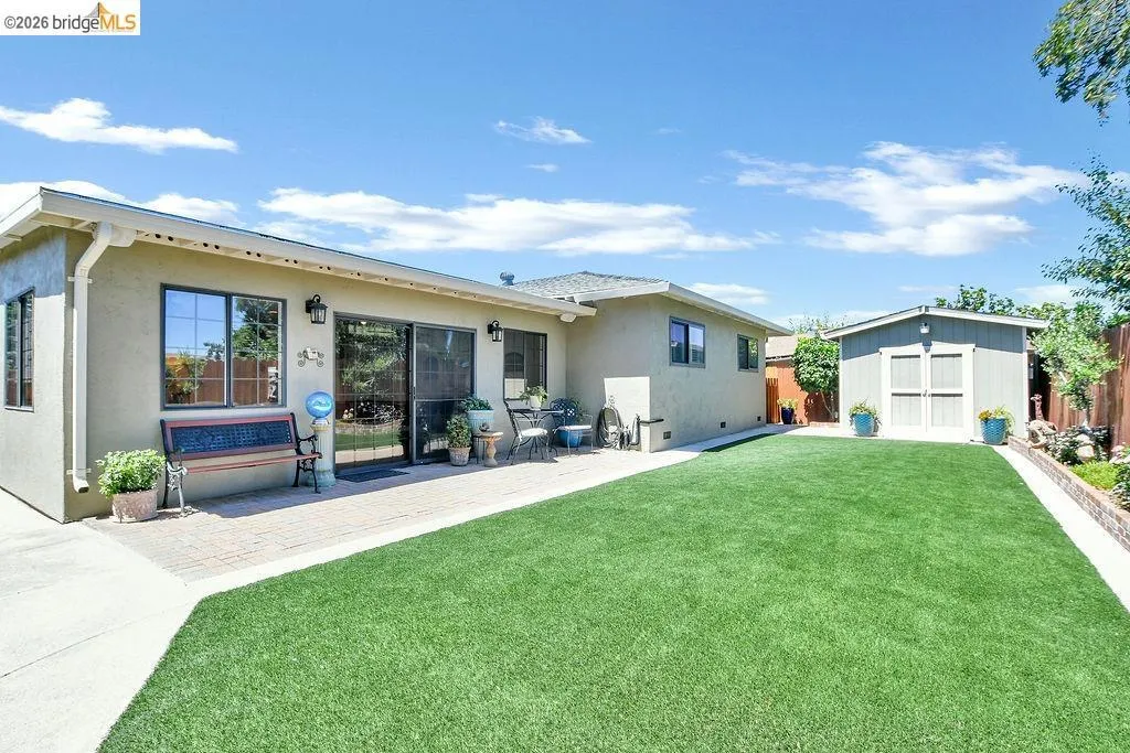 Expansive backyard featuring a lush green lawn, a paver patio, a concrete pathway, a detached storage shed with double doors, and a raised garden bed along the perimeter