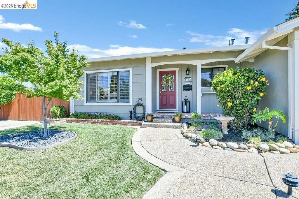 Single-story residence featuring a red entry door, an exposed aggregate pathway, a manicured lawn, an ornamental tree, and mature landscaping