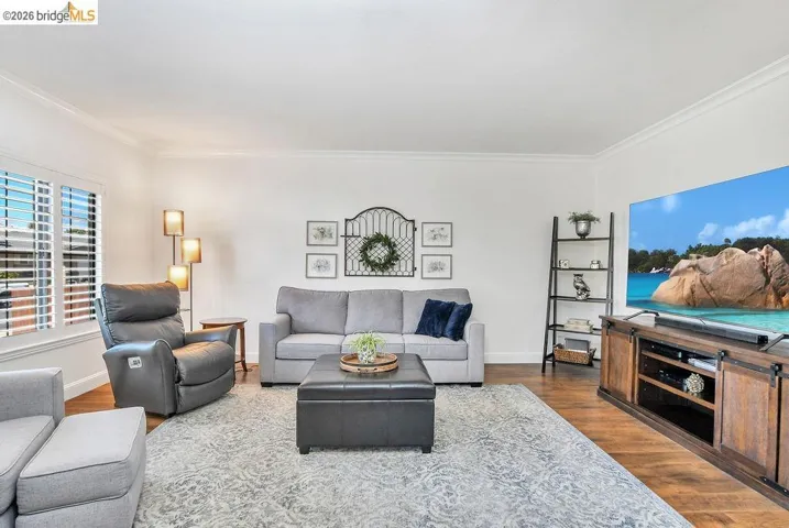 Spacious living area featuring crown molding, plantation shutters, wood-finish flooring, a decorative area rug, and a media console with barn door​​‌​​​​‌​​‌‌​​‌​​​‌‌​​​‌​​‌‌​​‌‌​​‌‌​​​​ accents