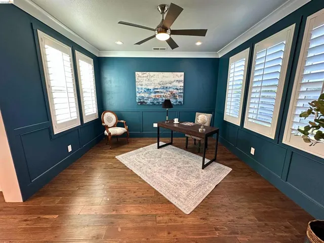 Office space featuring wood-finish flooring, deep blue walls with wainscoting, white plantation shutters, crown molding, and a ceiling fan with integrated lighting