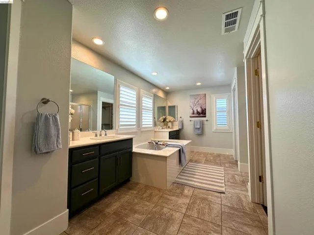 Spacious bathroom featuring an integrated soaking tub, dual vanities with dark cabinetry, plantation shutters, recessed lighting, and large format tile flooring