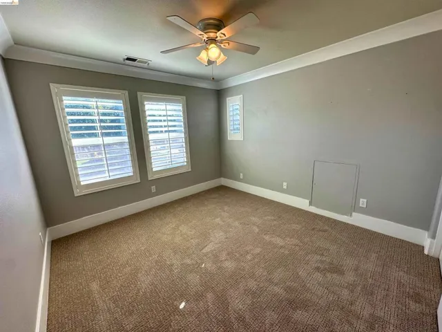 Spacious room with plush carpet flooring, two large windows with white louvered shutters, crown molding, and a ceiling fan with light fixture