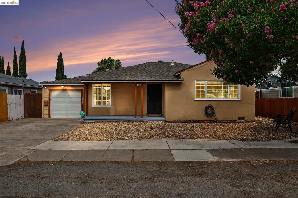 Single-story residence featuring a stucco exterior, attached garage with a roll-up door, covered front porch with wood columns, and a gravel front yard