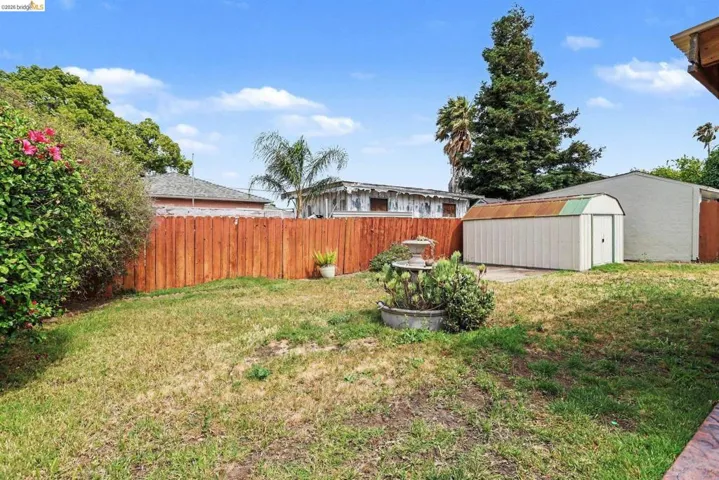 Expansive lawn area featuring a wooden fence, mature landscaping, a multi-tiered fountain, and a detached storage shed on a concrete pad