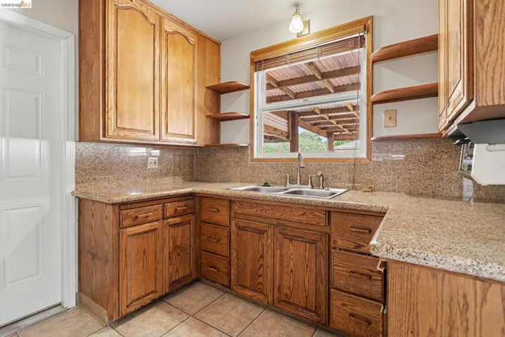 Kitchen featuring wood cabinetry with ample storage, a double basin stainless steel sink, natural stone countertops, a coordinated stone backsplash, and tile flooring