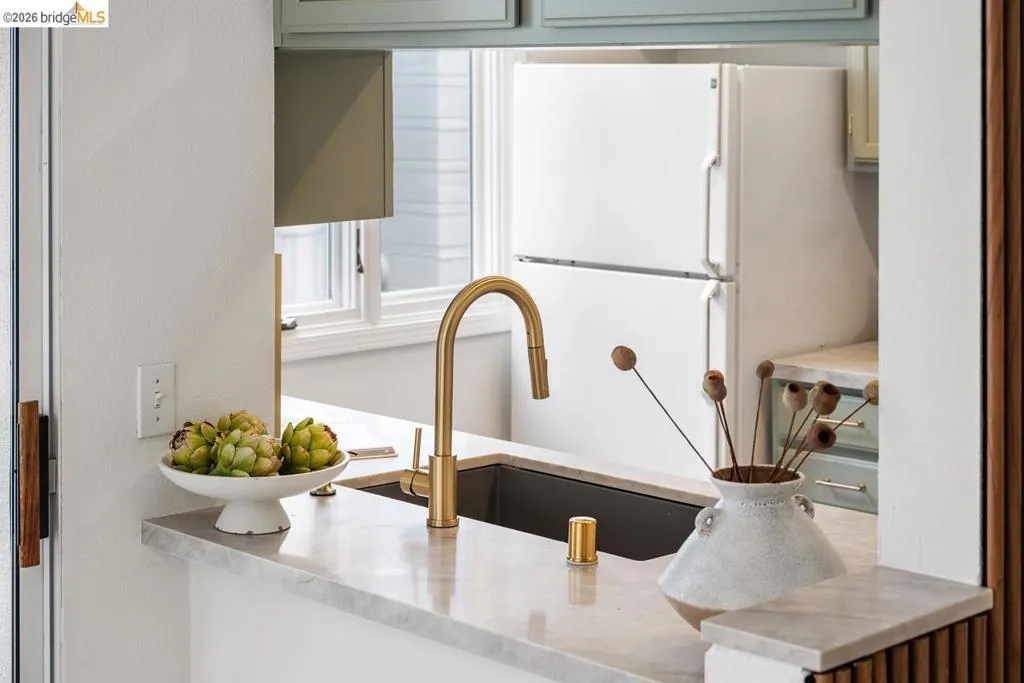 Kitchen pass-through featuring a deep-set sink with a brass-finish faucet, light-toned countertop, and light green cabinetry Kitchen pass-through featuring a deep-set sink with a brass-finish faucet, light-toned countertop, and light green cabinetry