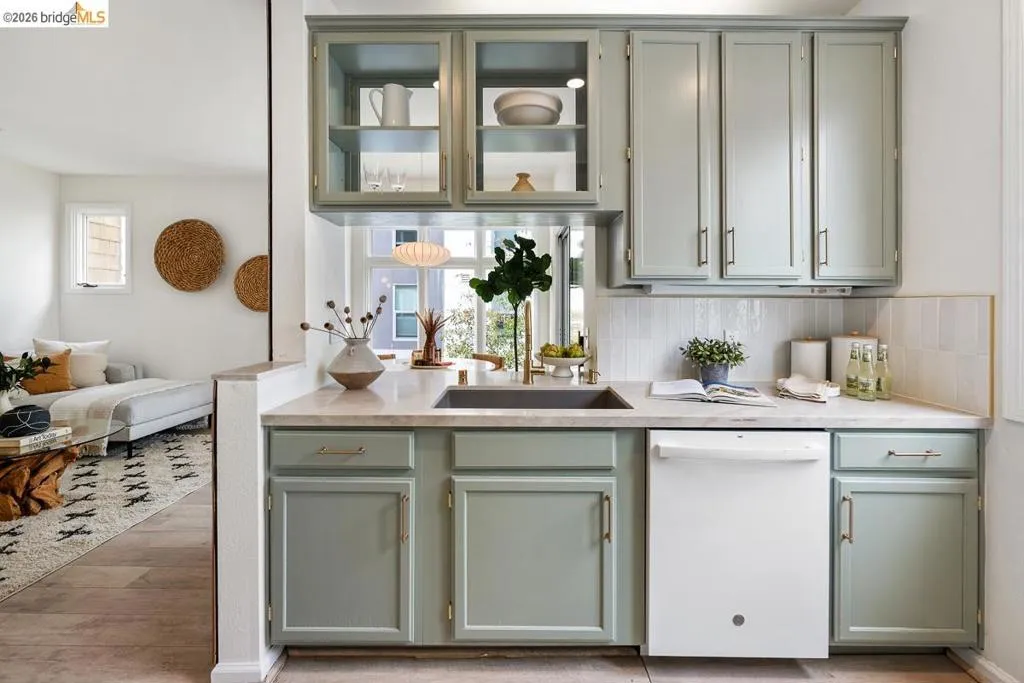 Kitchenette featuring light green cabinetry, clear glass upper cabinets, a built-in sink with a gold-tone faucet, and a white dishwasher Kitchenette featuring light green cabinetry, clear glass upper cabinets, a built-in sink with a gold-tone faucet, and a white dishwasher