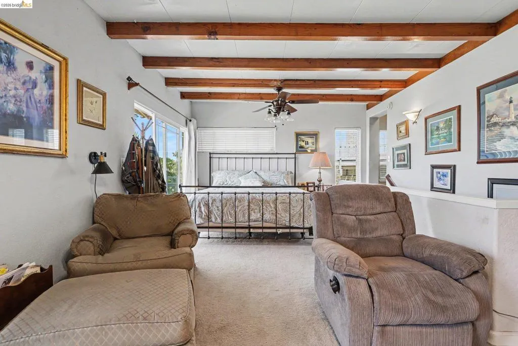 Spacious room featuring exposed wood ceiling beams, light-colored carpeting, and a ceiling fan with integrated lighting