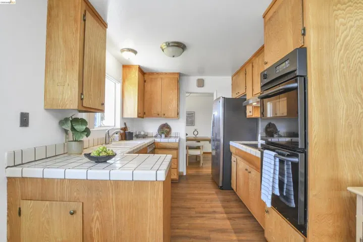 Kitchen featuring wood-finish cabinetry, tile countertops, built-in oven, stainless-steel refrigerator, and wood-finish flooring