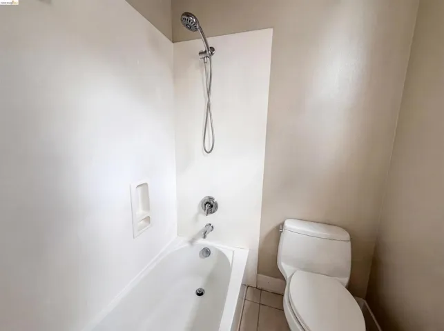 Bathroom featuring a white tub with integrated shelving, chrome shower fixtures, a handheld showerhead, and a white two-piece toilet