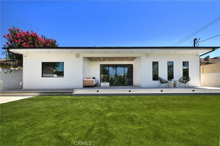 Full wide-angle view of the modern single-story exterior and expansive, flat grassy lawn, with an outdoor lounge space featuring a built-in surround sound system, ceiling heater