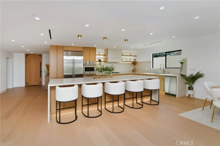 Gourmet kitchen featuring an oversized waterfall island, custom bar seating, warm wood cabinetry, open shelving with integrated lighting, and premium stainless steel appliances